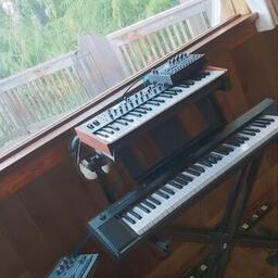 Synths, keyboard and sampler setup in front of a window giving into the forest